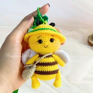 May include: A handmade crochet bee keyring. The bee is yellow with brown stripes, white wings, and a small beige satchel. It wears a yellow hat with green accents. The keyring is held by a hand.