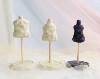 May include: Three crocheted mannequins on wooden stands. The mannequins are in different colors: white, cream, and purple. The stands are on a white surface.