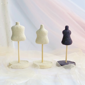 Op de afbeelding: Drie gehaakte mannequins op houten standaards. De mannequins zijn in verschillende kleuren: wit, crème en paars. De standaards staan op een wit oppervlak.