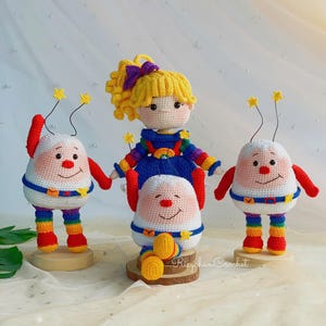 Könnte beinhalten: Eine Sammlung handgefertigter Häkelpuppen. Die Puppen zeigen ein Mädchen mit gelbem Haar und einer lila Schleife sowie drei skurrile Figuren mit roten und weißen Hüten und Outfits in Regenbogenfarben. Die Puppen werden auf Holzsockeln präsentiert.