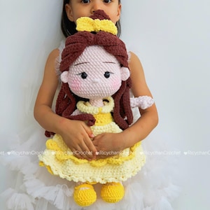 May include: A handmade crochet doll featuring long brown yarn hair styled with a yellow bow, a pale pink face with embroidered features, and a yellow dress with a ruffled skirt. The doll wears matching yellow shoes and has a soft, textured finish.
