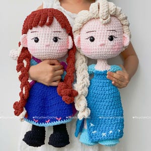 Op de afbeelding: Twee handgemaakte gehaakte poppen, een met roodbruine vlechten en een blauwe jurk, de andere met blonde vlechten en een blauwe jurk. Beide poppen hebben een roze huid en zwarte ogen. Ze worden vastgehouden door een persoon die een witte jurk draagt.