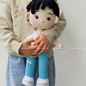 Puede incluir: Muñeco de ganchillo hecho a mano que representa a un niño. El muñeco tiene pelo negro, mejillas rosadas y viste una camisa blanca y pantalones azules. Los pies del muñeco son blancos con un ribete beige. El muñeco está siendo sujetado.