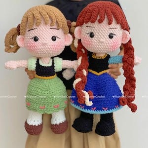 May include: Two handmade crochet dolls, one with light brown pigtails and a green dress, the other with red braids and a blue dress. Both dolls have rosy cheeks and black eyes. The dolls are held up against a neutral background.