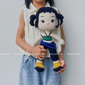 Puede incluir: Muñeca de ganchillo hecha a mano con cabello negro recogido en dos moños. La muñeca tiene una cara color crema con mejillas rosadas y viste un atuendo verde y azul con detalles amarillos. La muñeca es sostenida por una persona.
