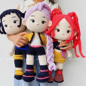 Puede incluir: Tres muñecas de ganchillo hechas a mano con detalles. Cada muñeca tiene un peinado y un atuendo únicos, incluyendo una muñeca con una trenza morada, una muñeca con coletas rojas y una muñeca con cabello negro. Las muñecas se sostienen frente a un fondo claro.