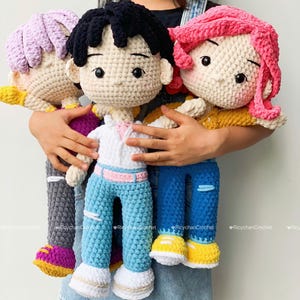 Puede incluir: En la imagen se sostienen tres muñecas de ganchillo hechas a mano. Cada muñeca tiene un peinado y un atuendo únicos. Una muñeca tiene cabello negro y vaqueros azules, otra tiene cabello rosa y vaqueros azules, y la tercera tiene cabello morado y pantalones grises. Las muñecas tienen zapatos amarillos y blancos.