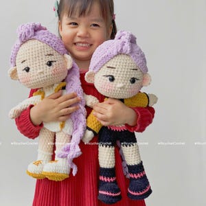 May include: A young girl holding two handmade crochet dolls. The dolls have light skin, purple hair, and are wearing outfits with yellow, black, and white accents. The girl is wearing a red dress. The dolls are handmade.