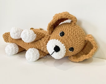 May include: A crocheted brown and white dog plush toy with black eyes and a black nose. The toy is lying on its side with its head resting on the ground.