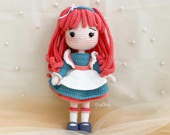 Puede incluir: Una muñeca de crochet con el pelo largo y rojo, vestida con un vestido azul y blanco con un delantal blanco. La muñeca tiene ojos negros y una cara rosa.
