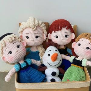 Könnte beinhalten: Eine Sammlung von fünf handgefertigten Häkelpuppen mit Garnhaaren, gestickten Gesichtszügen und farbenfrohen Outfits, einschließlich eines Schneemanns, die in einer beigen Aufbewahrungsbox mit dem Aufdruck RicychanCrochet untergebracht sind.