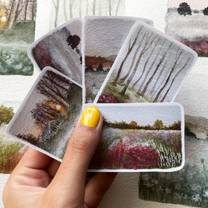 May include: A collection of rectangular watercolor paintings depicting serene landscape scenes. The paintings feature trees, fields, and buildings in muted tones of gray, brown, and purple. The artwork is held in a hand with yellow nail polish.