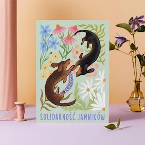 May include: A green background with two dachshunds playing in a field of flowers. The text "Solidarność Jamników" is at the bottom of the image.
