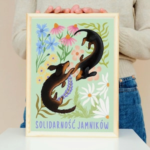 May include: A green background with two black and brown dachshunds playing in a field of colourful flowers. The text "Solidarność Jamników" is at the bottom of the image.