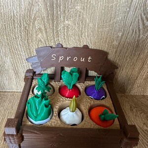 May include: A brown wooden toy garden box with the word "Sprout" on a sign. The box contains six colorful toy vegetables: green lettuce, a red radish, a purple turnip, a green cabbage, a white garlic, and an orange tomato.