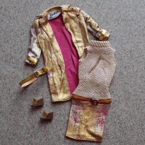May include: A vintage doll outfit featuring a gold metallic coat with a pink lining, a matching gold skirt and top, a gold belt, and brown shoes. The coat has a label that reads "Barbie by Mattel".