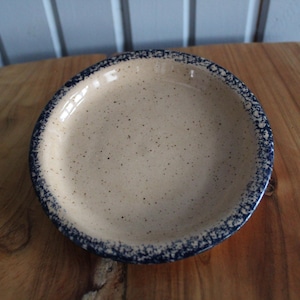 May include: A small, round, ceramic dish with a speckled beige glaze and a blue rim. The dish has a speckled blue and white design around the edge.