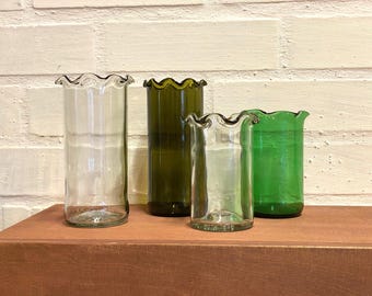 May include: Four glass vases with ruffled tops. The vases are clear, green, and brown. They are arranged on a wooden surface.