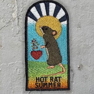 Pode incluir: Patch bordado com um rato castanho em frente a um sol amarelo e céu azul. Um coração vermelho e o texto "HOT RAT SUMMER" também estão incluídos. O patch tem uma borda preta. Fundo cinza.
