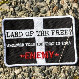 Może przedstawiać: Prostokątna czarno-biała naszywka z napisem "LAND OF THE FREE?" i "WHOEVER TOLD YOU THAT IS YOUR ENEMY" w kolorze białym i czerwonym. Naszywka ma biały wzór krzyża i jest umieszczona na tle kamyków i mchu.