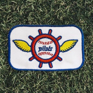 May include: A rectangular patch with a white background and a blue border. The patch features a red baseball-themed design with the word "pilots" in the center, surrounded by a red wheel and yellow wings. The patch is set against a green grass background.