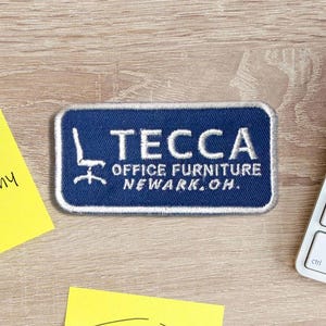 May include: A navy blue patch with white embroidered text that reads "TECCA OFFICE FURNITURE NEWARK, OH." A small white chair graphic is on the left. Two yellow sticky notes are also visible, one with the word "company" and the other with the words "red ball market global."