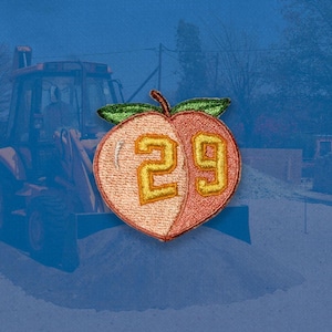 Puede incluir: Parche bordado en forma de melocotón con el número 29 en dorado. El melocotón está dividido en dos colores, con un color melocotón claro a la izquierda y un color naranja rojizo a la derecha. Hojas verdes y un tallo marrón en la parte superior.