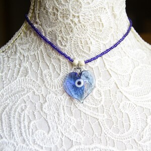 May include: A blue glass heart pendant necklace with an evil eye charm. The necklace is made of small blue beads and has a white bead at the top.