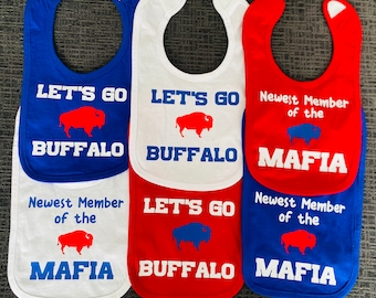 Puede incluir: Tres baberos para bebé con diferentes diseños. El primer babero es azul con texto blanco que dice "Let's Go Buffalo" y una silueta de búfalo roja. El segundo babero es blanco con texto azul que dice "Let's Go Buffalo" y una silueta de búfalo roja. El tercer babero es rojo con texto blanco que dice "Newest Member of the Mafia" y una silueta de búfalo azul.