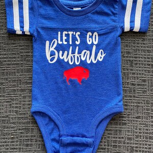 May include: A blue baby onesie with white stripes on the sleeves. The onesie has the text "Let's Go Buffalo" in white with a red buffalo silhouette.
