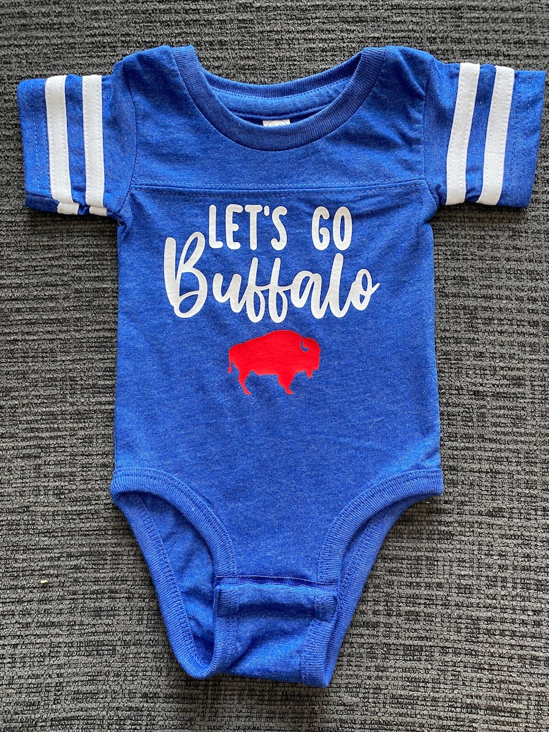 May include: A blue baby onesie with white stripes on the sleeves. The onesie has the text "Let's Go Buffalo" in white with a red buffalo silhouette.