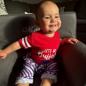 May include: A baby boy wearing a red shirt with the text "Let's Go Buffalo" in white. He is wearing blue and white striped pants and is sitting in a gray armchair.