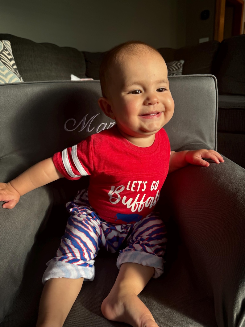 May include: A baby boy wearing a red shirt with the text "Let's Go Buffalo" in white. He is wearing blue and white striped pants and is sitting in a gray armchair.