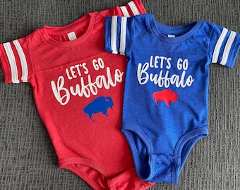 Puede incluir: Dos bodies de bebé, uno rojo y otro azul, con rayas blancas en las mangas. Los bodies tienen el texto "Let's Go Buffalo" con una silueta de búfalo azul en el body rojo y una silueta de búfalo rojo en el body azul.
