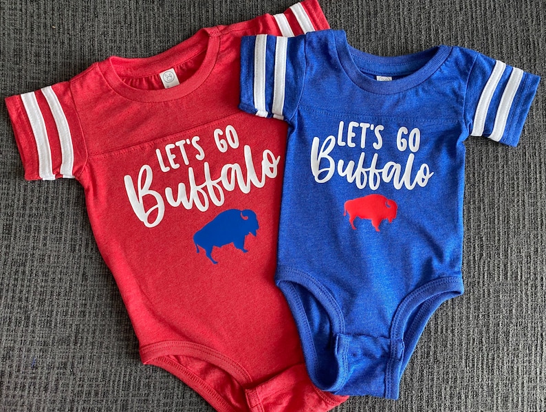 May include: Two red and blue baby onesies with white stripes on the sleeves. The onesies have the text "Let's Go Buffalo" with a blue buffalo silhouette on the red onesie and a red buffalo silhouette on the blue onesie.