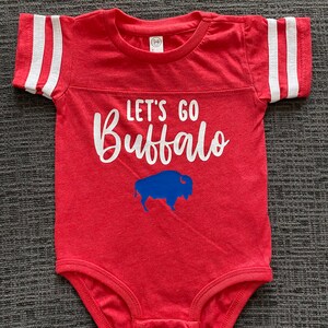 May include: Red baby onesie with white stripes on the sleeves. The onesie has the text "Let's Go Buffalo" in white with a blue buffalo silhouette below the text.