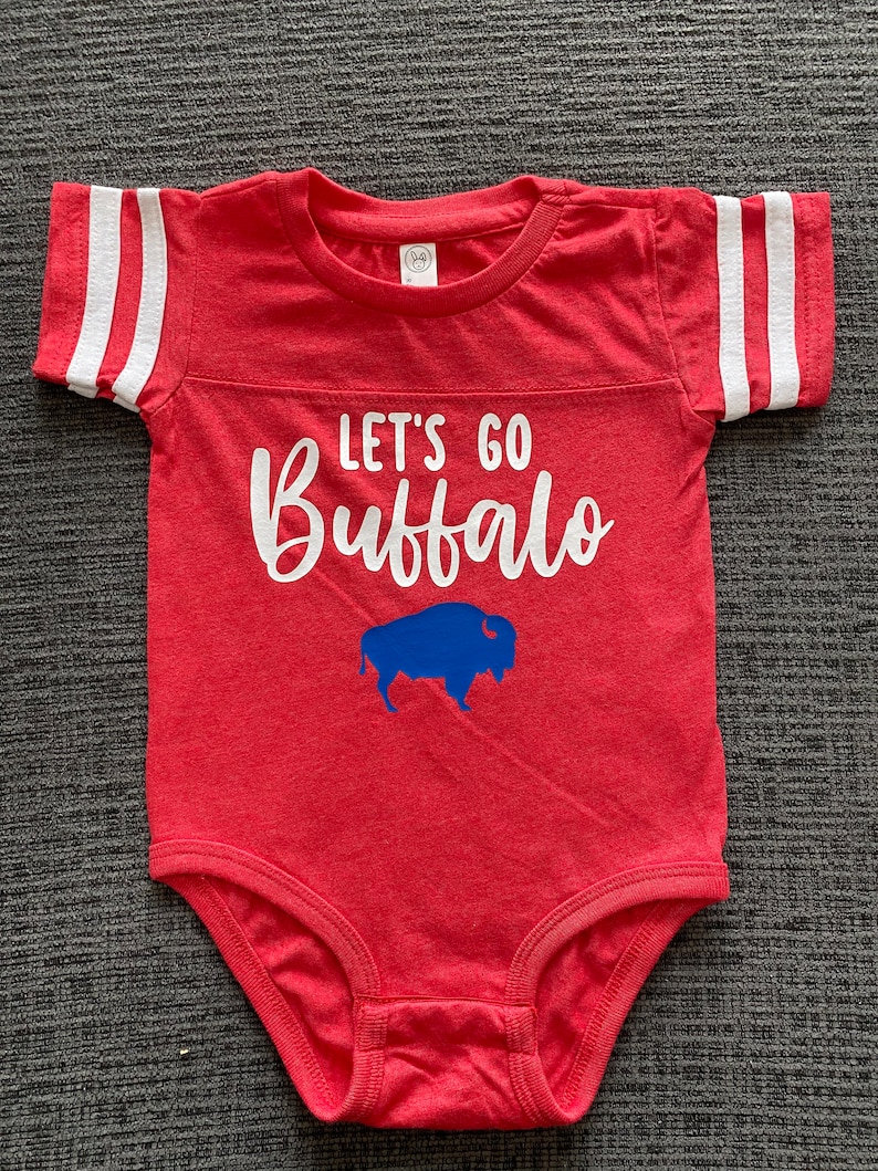 May include: Red baby onesie with white stripes on the sleeves. The onesie has the text "Let's Go Buffalo" in white with a blue buffalo silhouette below the text.