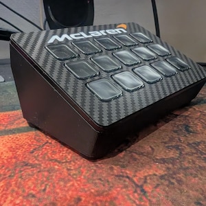 May include: A black, carbon fibre button box with a McLaren logo and 16 buttons. The buttons are clear and have a white outline.