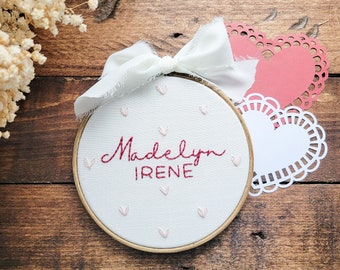 Puede incluir: Un aro de bordado blanco con el nombre "Madelyn Irene" bordado en hilo rojo y corazones rosas. El aro está decorado con un lazo de cinta blanco y dos servilletas de papel, una rosa y otra blanca.
