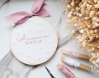 May include: A white embroidery hoop with the name "Arianne Maria" embroidered in pink thread. The hoop is surrounded by white and pink embroidery floss on wooden clothespins and a pair of silver embroidery scissors.
