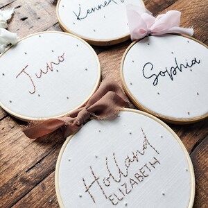 Puede incluir: Cuatro aros de bordado blancos con marcos dorados, cada uno con un nombre diferente bordado en hilo negro o marr&oacute;n. Los nombres son "Kennedy", "Juno", "Sophia" y "Holland Elizabeth".