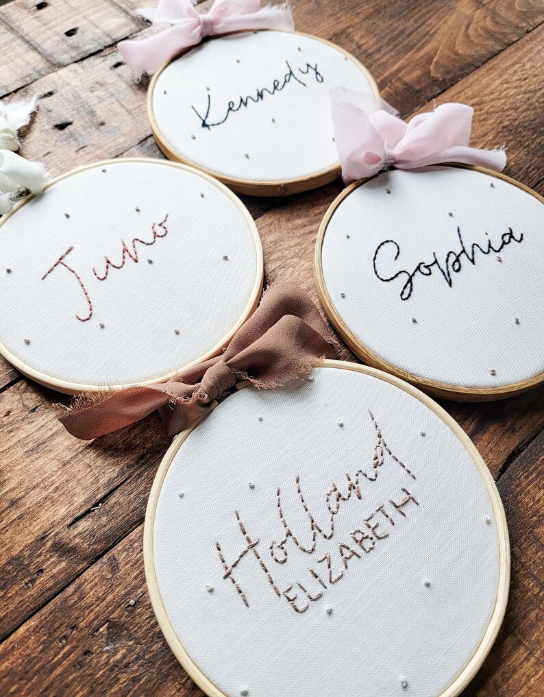 Puede incluir: Cuatro aros de bordado blancos con marcos dorados, cada uno con un nombre diferente bordado en hilo negro o marr&oacute;n. Los nombres son "Kennedy", "Juno", "Sophia" y "Holland Elizabeth".