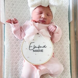 Puede incluir: Un pelele rosa para beb&eacute; con una diadema blanca con lazo y un aro de madera bordado con el nombre "Emma Marie".