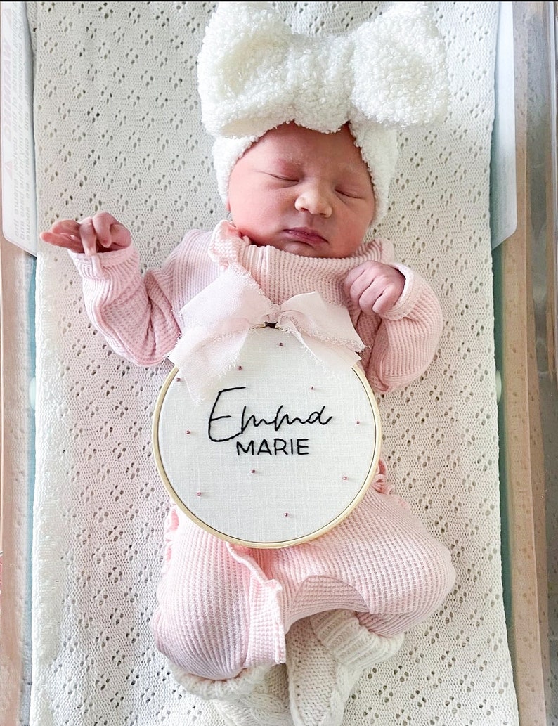 Puede incluir: Un pelele rosa para beb&eacute; con una diadema blanca con lazo y un aro de madera bordado con el nombre "Emma Marie".