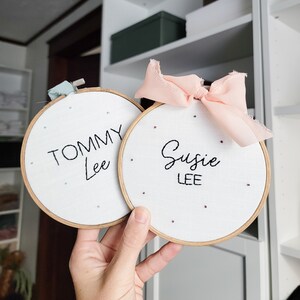 Puede incluir: Dos aros de bordado blancos con marcos de madera. El aro de la izquierda tiene el nombre "Tommy Lee" bordado en hilo negro. El aro de la derecha tiene el nombre "Susie Lee" bordado en hilo negro. Ambos aros tienen una cinta rosa y blanca atada alrededor.