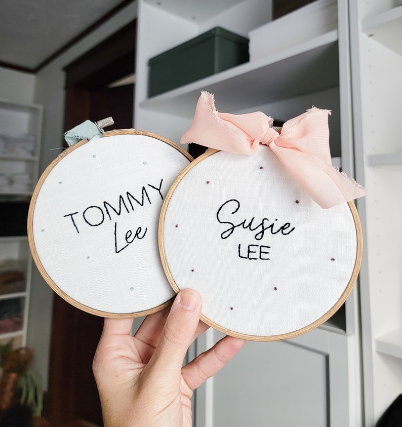 Puede incluir: Dos aros de bordado blancos con marcos de madera. El aro de la izquierda tiene el nombre "Tommy Lee" bordado en hilo negro. El aro de la derecha tiene el nombre "Susie Lee" bordado en hilo negro. Ambos aros tienen una cinta rosa y blanca atada alrededor.