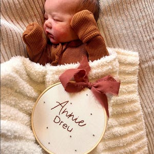 Puede incluir: Un aro blanco bordado con el nombre "Annie Drew" en hilo rojo. El aro est&aacute; atado con una cinta burdeos y est&aacute; sobre una manta blanca y peluda. Un beb&eacute; est&aacute; durmiendo en la manta con un mono de punto de waffle marr&oacute;n.