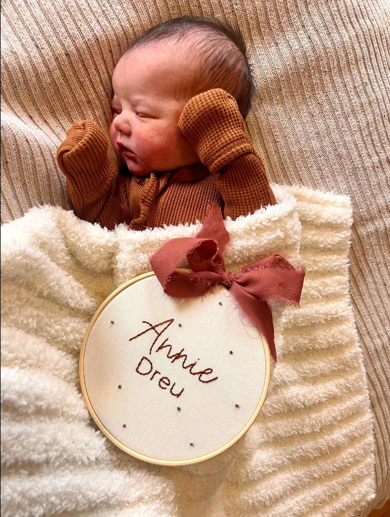 Puede incluir: Un aro blanco bordado con el nombre "Annie Drew" en hilo rojo. El aro est&aacute; atado con una cinta burdeos y est&aacute; sobre una manta blanca y peluda. Un beb&eacute; est&aacute; durmiendo en la manta con un mono de punto de waffle marr&oacute;n.