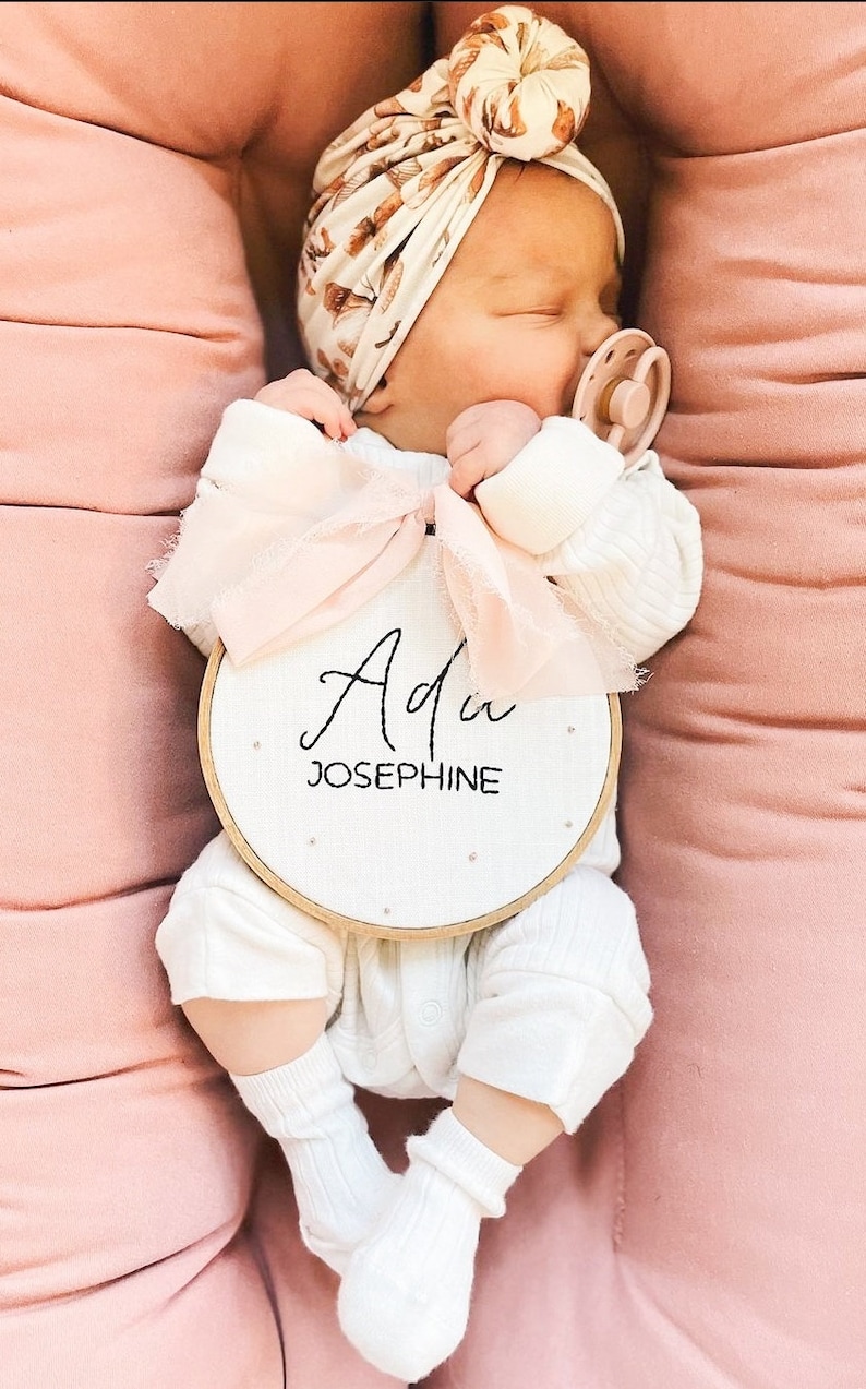Puede incluir: Un pelele blanco para beb&eacute; con un lazo rosa y un aro de bordado de madera con el nombre "Ada Josephine" bordado en hilo negro.