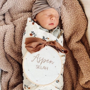 Puede incluir: Un beb&eacute; reci&eacute;n nacido con un turbante marr&oacute;n y un arrullo blanco con estampado floral. El beb&eacute; est&aacute; acostado sobre una manta peluda marr&oacute;n. Un aro de bordado de madera con tela blanca y el texto "Aspen Selah" est&aacute; delante del beb&eacute;.